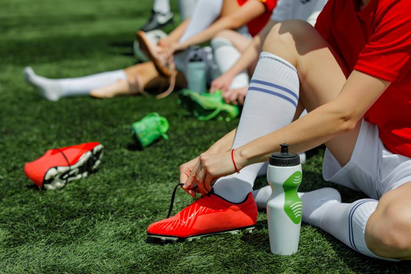 a youth male soccer player putting a soccer cleat on his foot while sitting on grass