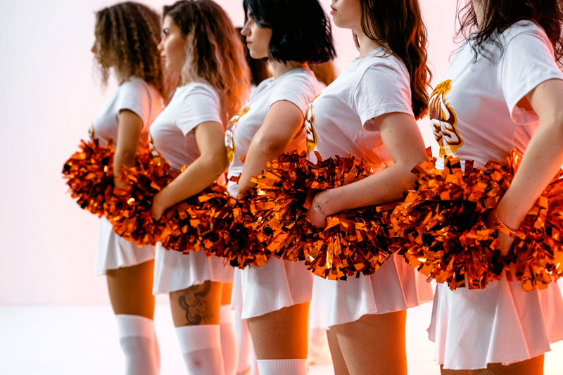 a squad of cheerleaders in white uniforms holding orange pom poms