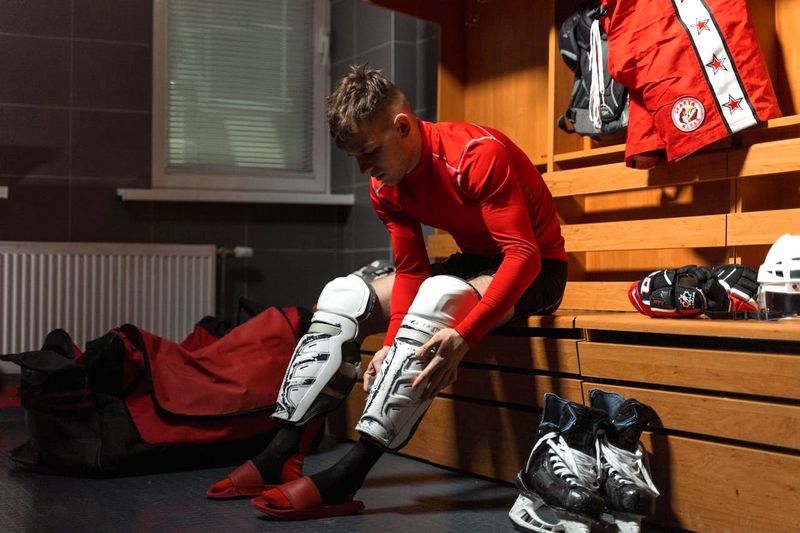 a young male ice hockey player putting on shin guards in a locker room