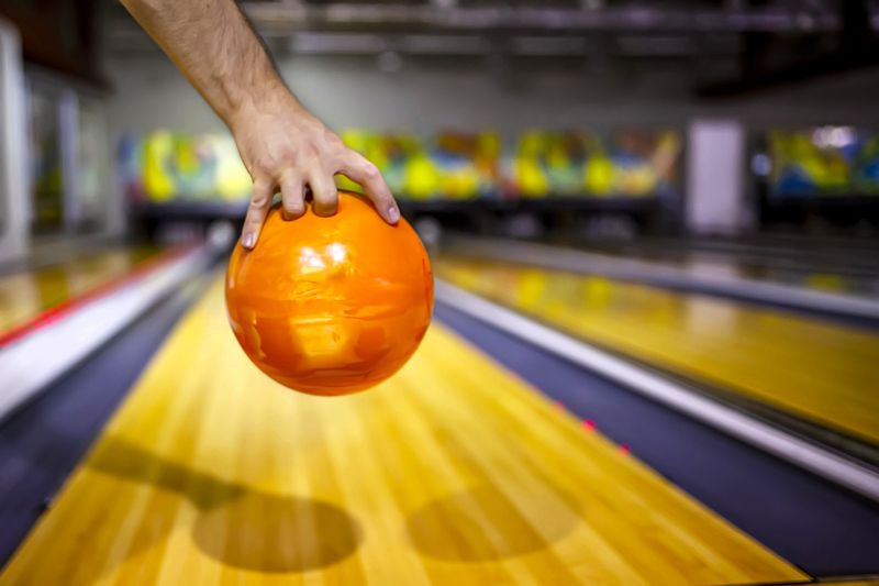 a hand holding an orange bowling ball in front a bowling lane