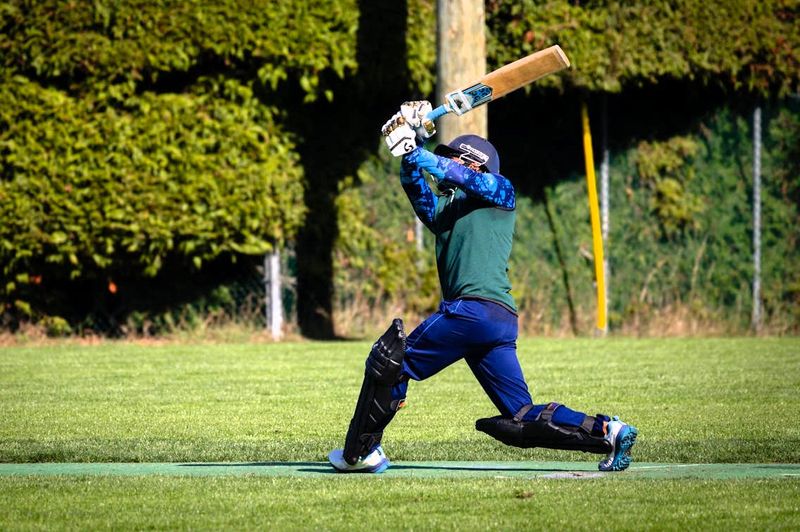 an adult cricket player swinging a cricket bat on a green grass pitch