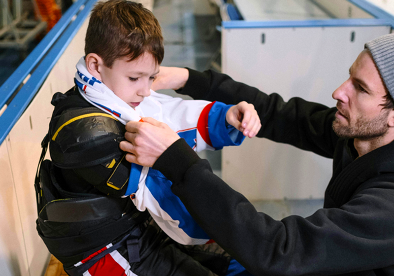 an adult male helping a youth boy ice hockey player put on his jersey next to an ice rink