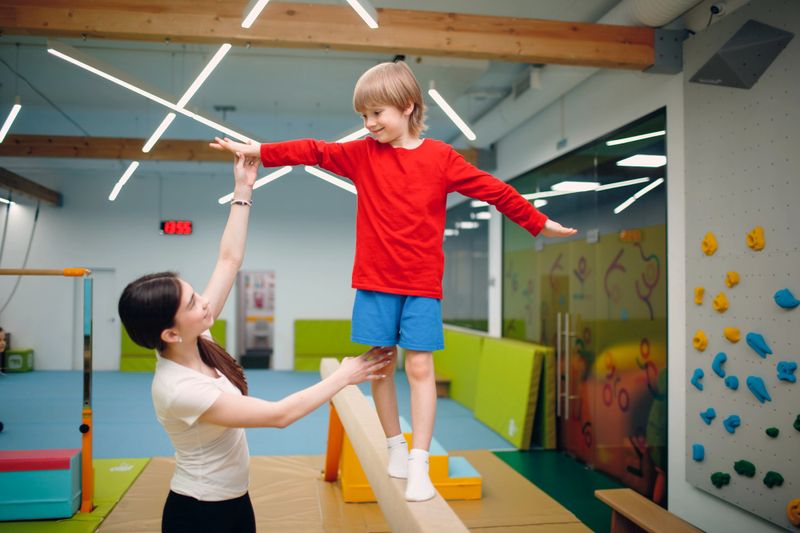 a young boy walking along a gymnastics beam with support of a gymnastics coach to the right of the child