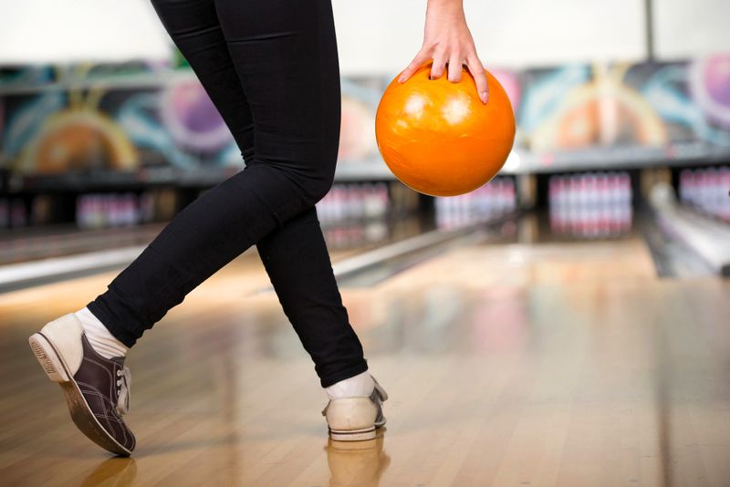 an adult female holding an orange bowling bowl in front of a bowling lane
